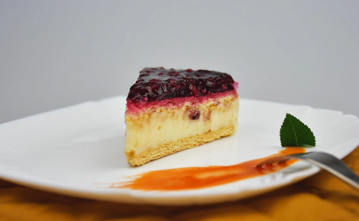 Trójkątny kawałek sernika jaglanego z warstwą owocową na białym talerzu, obok dekoracja z listkiem i musem